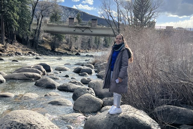 Daniela poses on a rock beside a river, with a bridge and mountains behind her in Colorado.