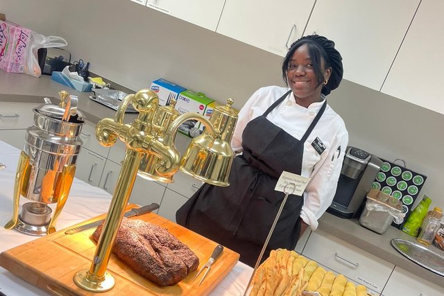 Katherine poses in a kitchen uniform behind a serving table with meat and bread.