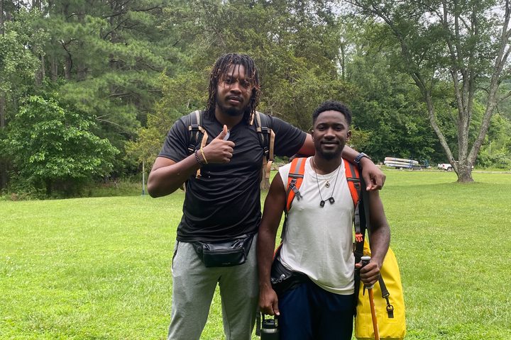 Dos jóvenes con mochilas y equipo de senderismo en un entorno arbolado, Summer Camp USA.